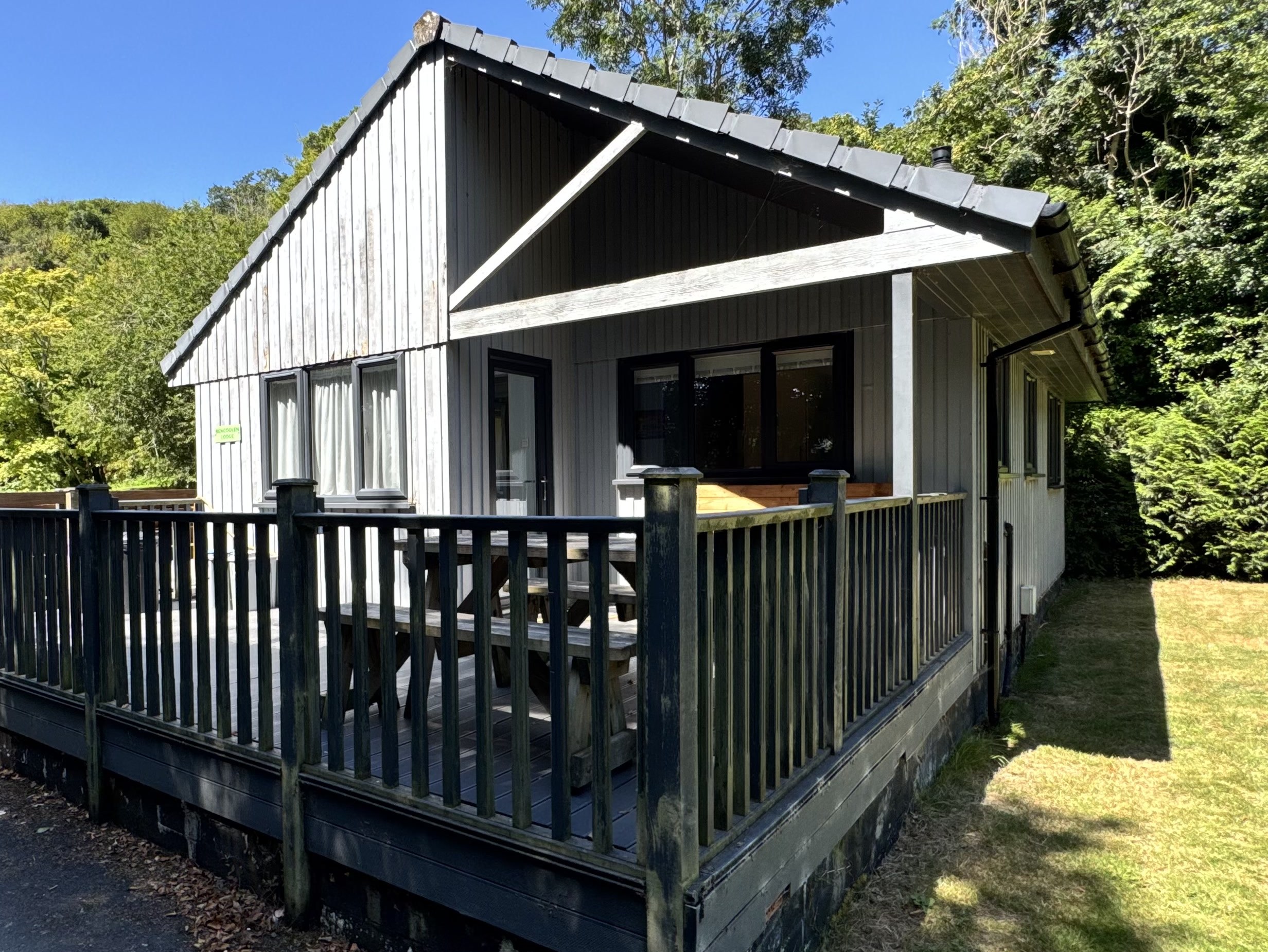 Bencoolen from outside: the timber lodge with its grey gable end and covered veranda, standing on a raised deck with a balustrade and a picnic table, the grass and the trees of the valley either side