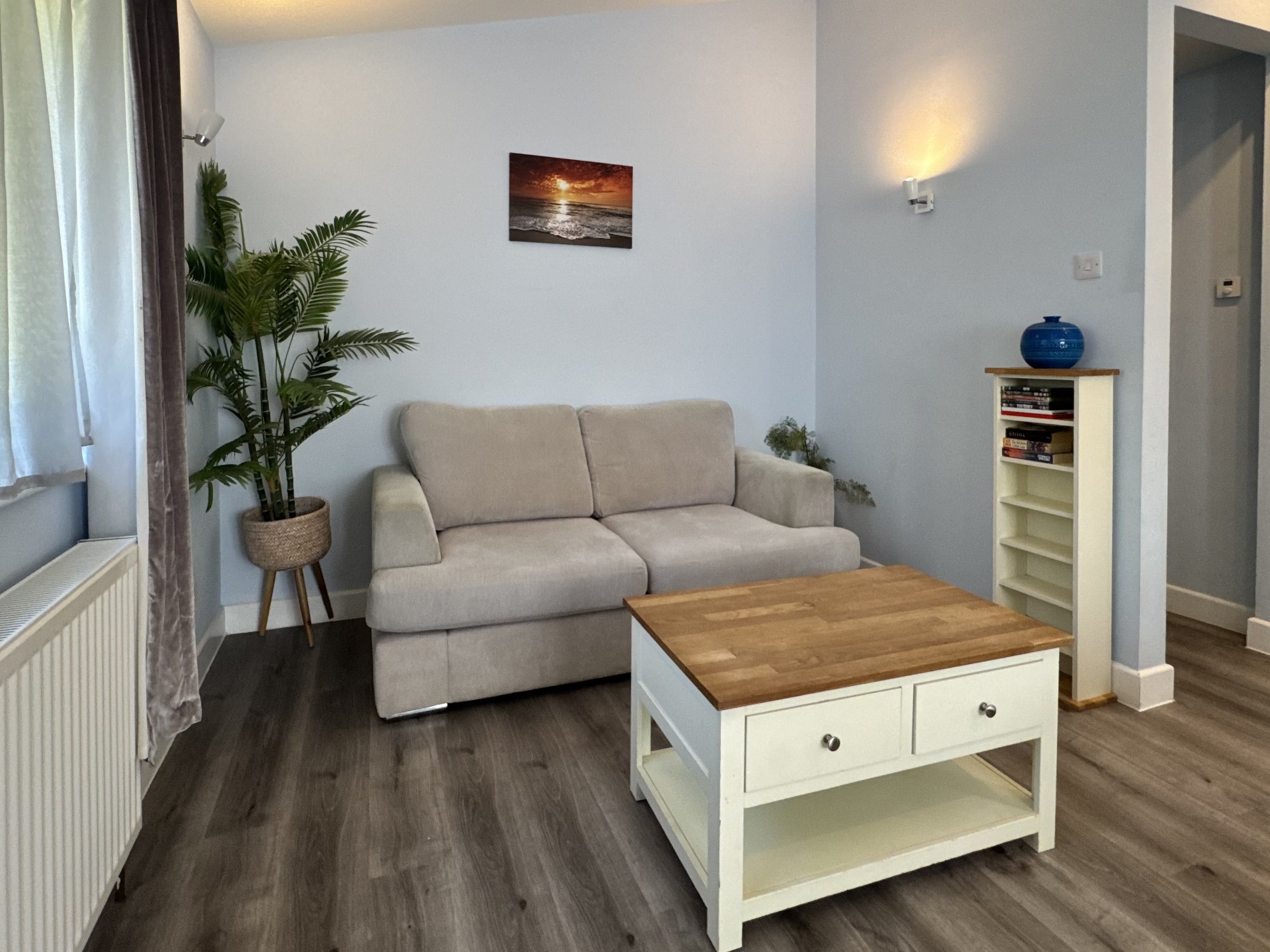 A corner of the lounge at Bencoolen, with a grey sofa, a potted palm, a sunset print on the wall and an oak-topped coffee table in front