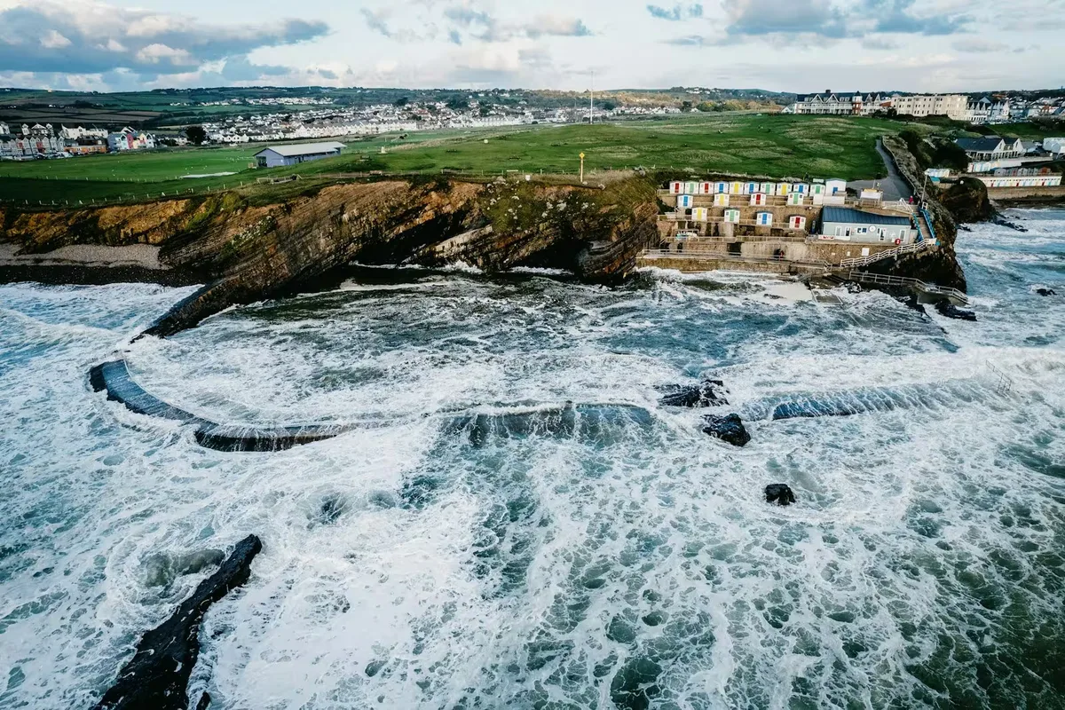 The sea pool and rock ledges at Bude from the air, with the beach huts, the clifftop field and the town behind