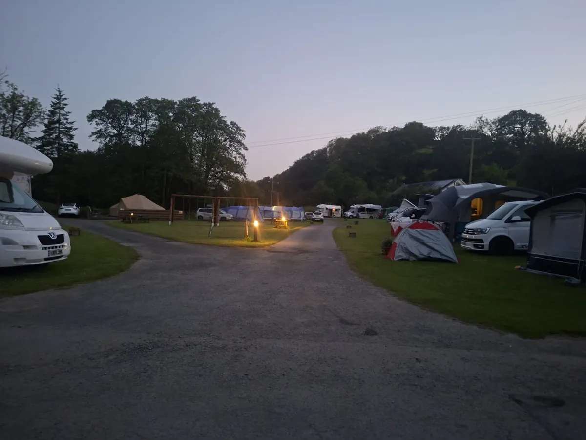 The camping field at dusk, the swings and picnic benches on the grass beyond the track, with tents and motorhomes pitched either side