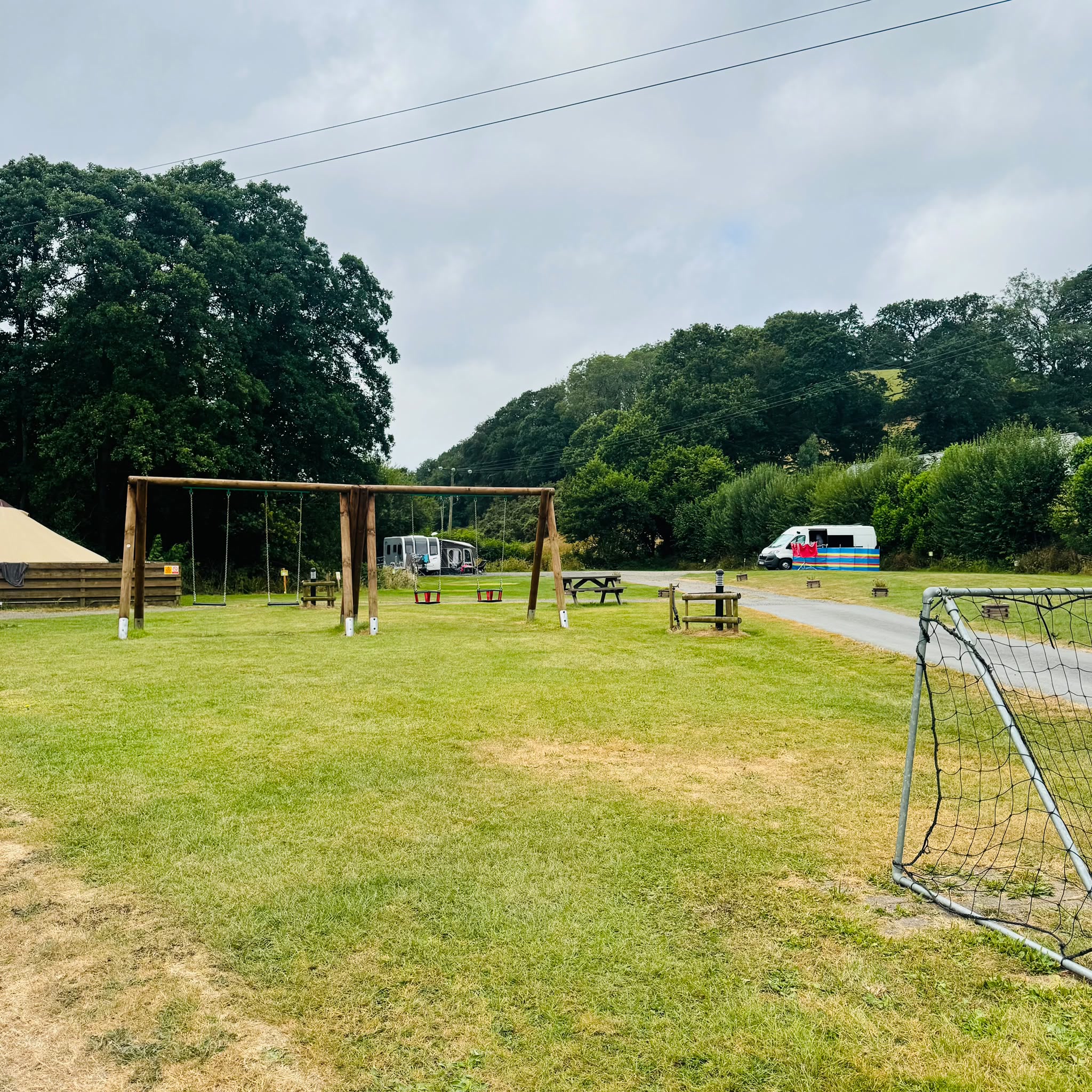 The swings and a football goal on the grass of the camping field, with picnic benches, a caravan and a campervan beyond and the wooded valley side behind