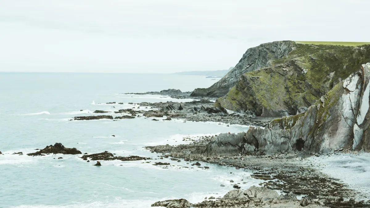 Grey slate cliffs and wave-cut rock ledges running along the North Cornwall coast under a pale sky, with white surf breaking over the reefs below
