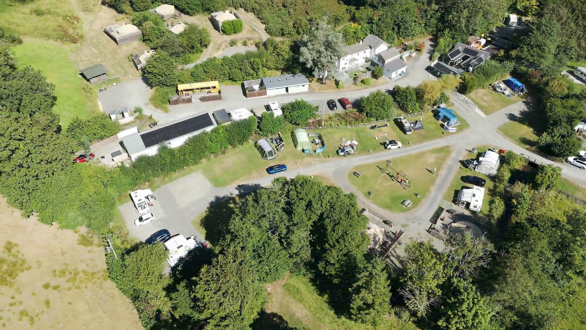 An aerial view over the park in summer: the camping field with tents and caravans, the swings on the grass triangle, the yellow school bus by the lane, and bell tents on their decks on the hillside above