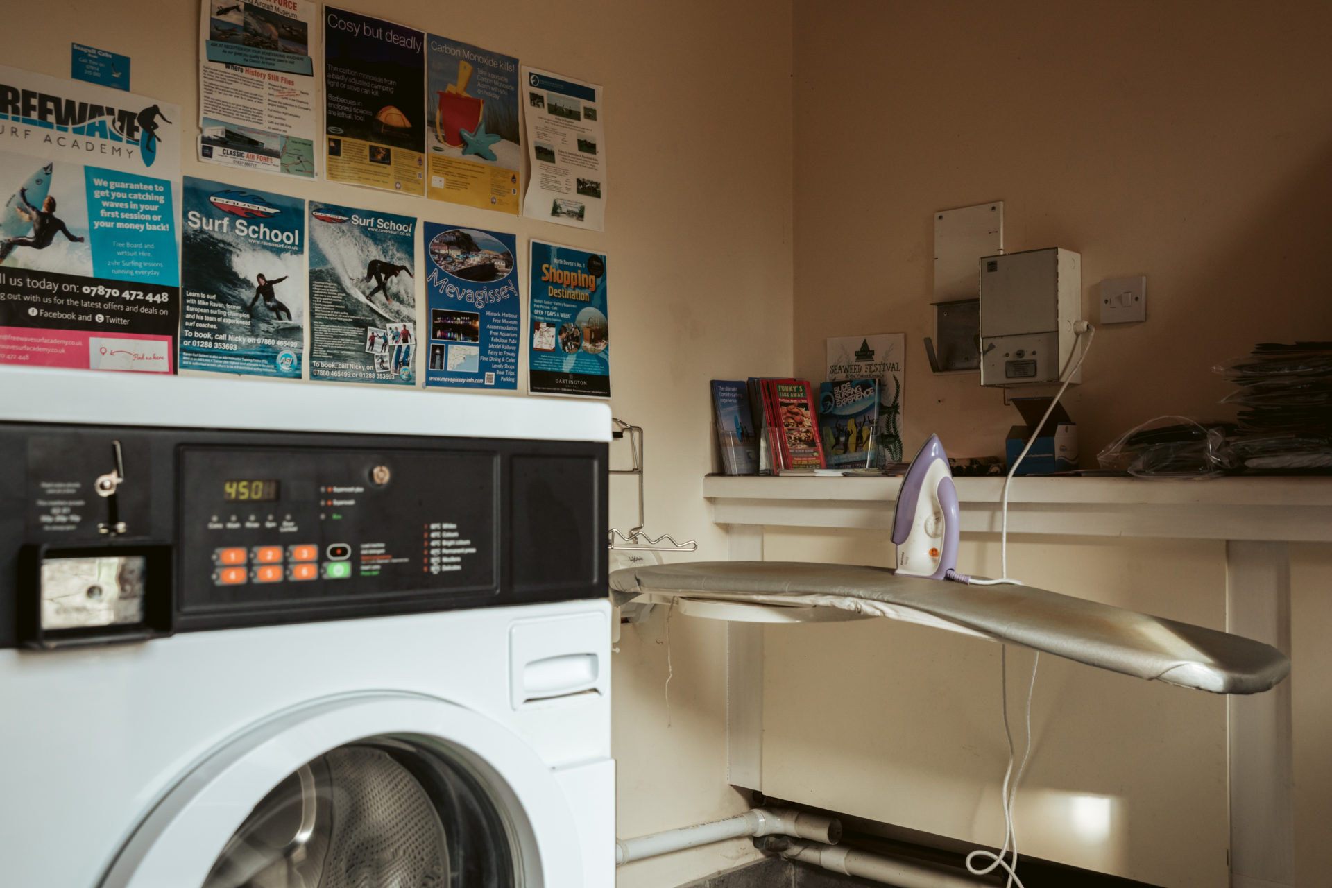 Inside the launderette: a washing machine with its coin meter alongside and an ironing board set up beside it