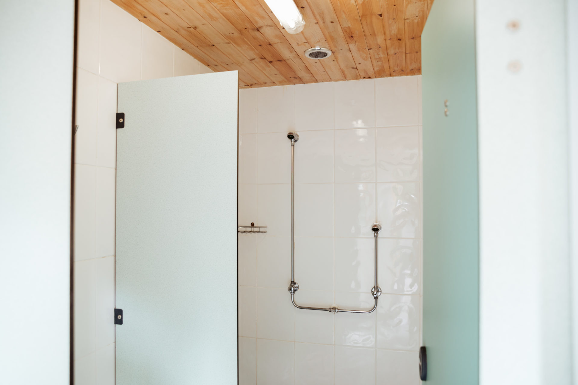 A shower cubicle in the shared block, tiled white with a frosted privacy door and a pine-boarded ceiling above