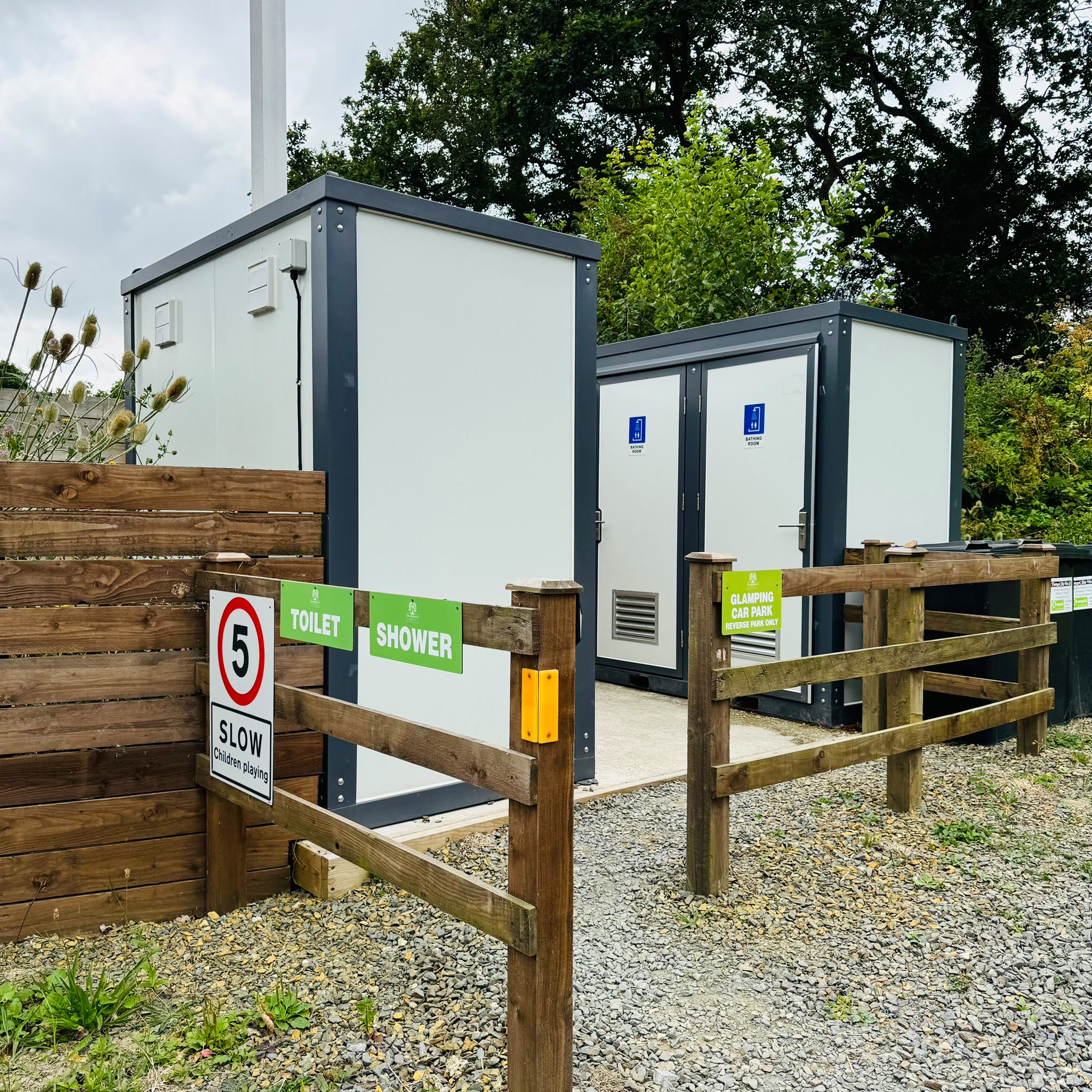 The toilet and shower pods in the glamping car park: two modern white units behind a timber rail with green TOILET and SHOWER signs