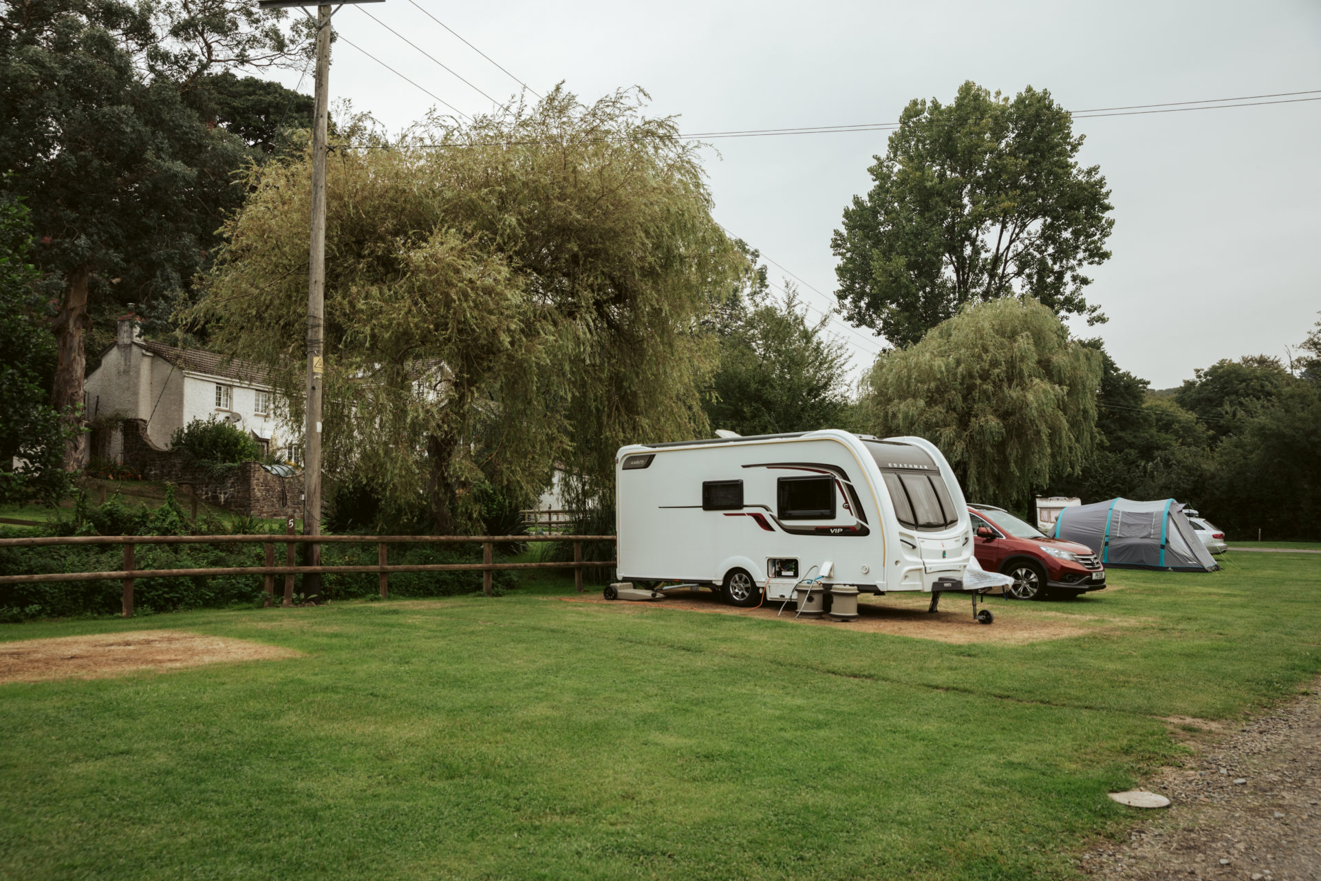 A caravan with a car alongside and a family tent on the next pitch, on level grass with willows and the valley side behind