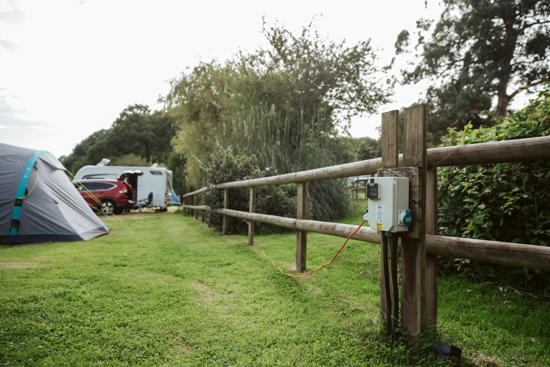 A level grass pitch mown short, its electric hook-up post on the post-and-rail fence with the cable run out, a tent to one side and a caravan and car at the top of the field