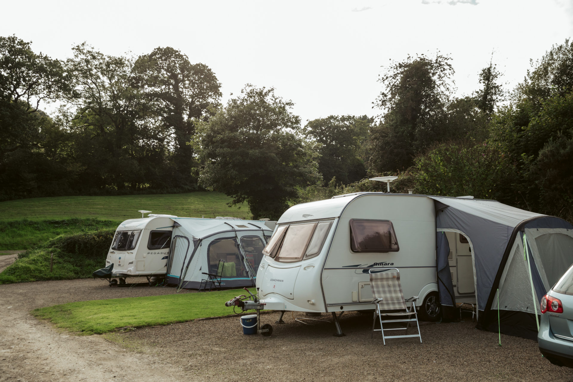 Two caravans with awnings on the gravel hardstandings, a folding chair out beside one of them and the trees and a green field rising behind
