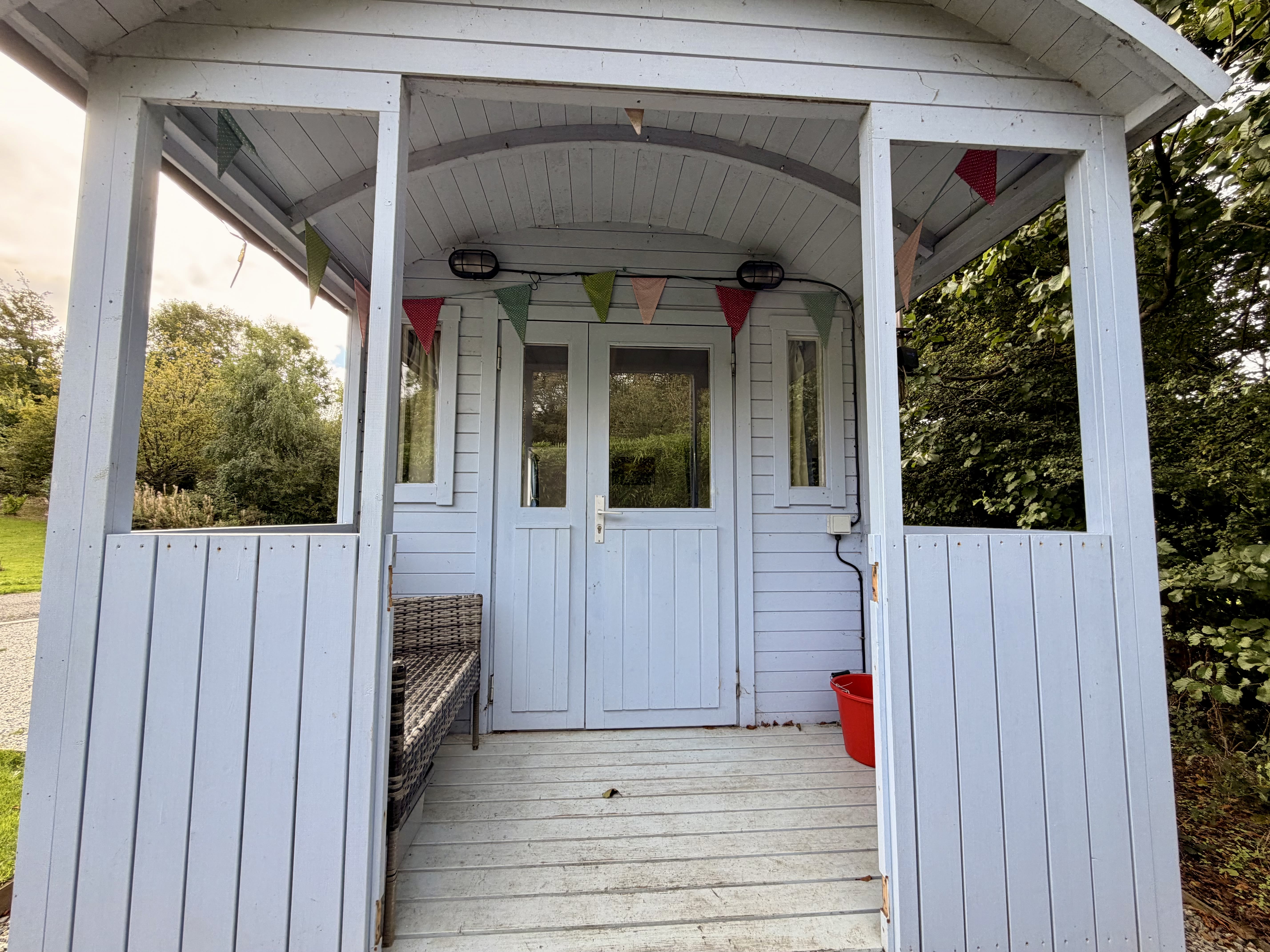 Millook's covered porch, pale blue-grey boarding with bunting strung overhead, a rattan bench and the glazed stable door into the hut