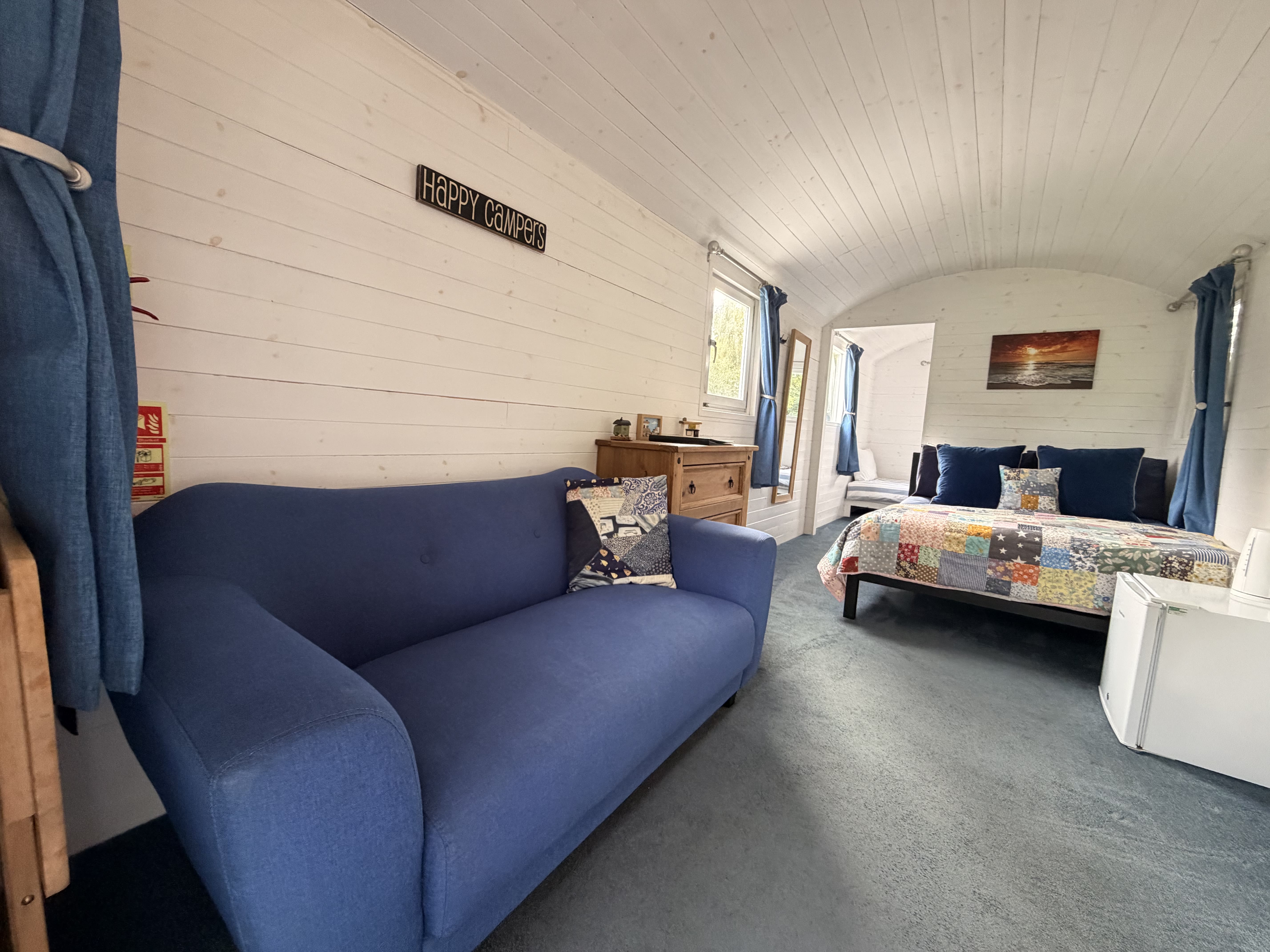 Looking down the hut from the blue sofa past a chest of drawers to the patchwork-quilted bed at the far end, with the "Happy Campers" sign on the wall and a sunset print above the bed