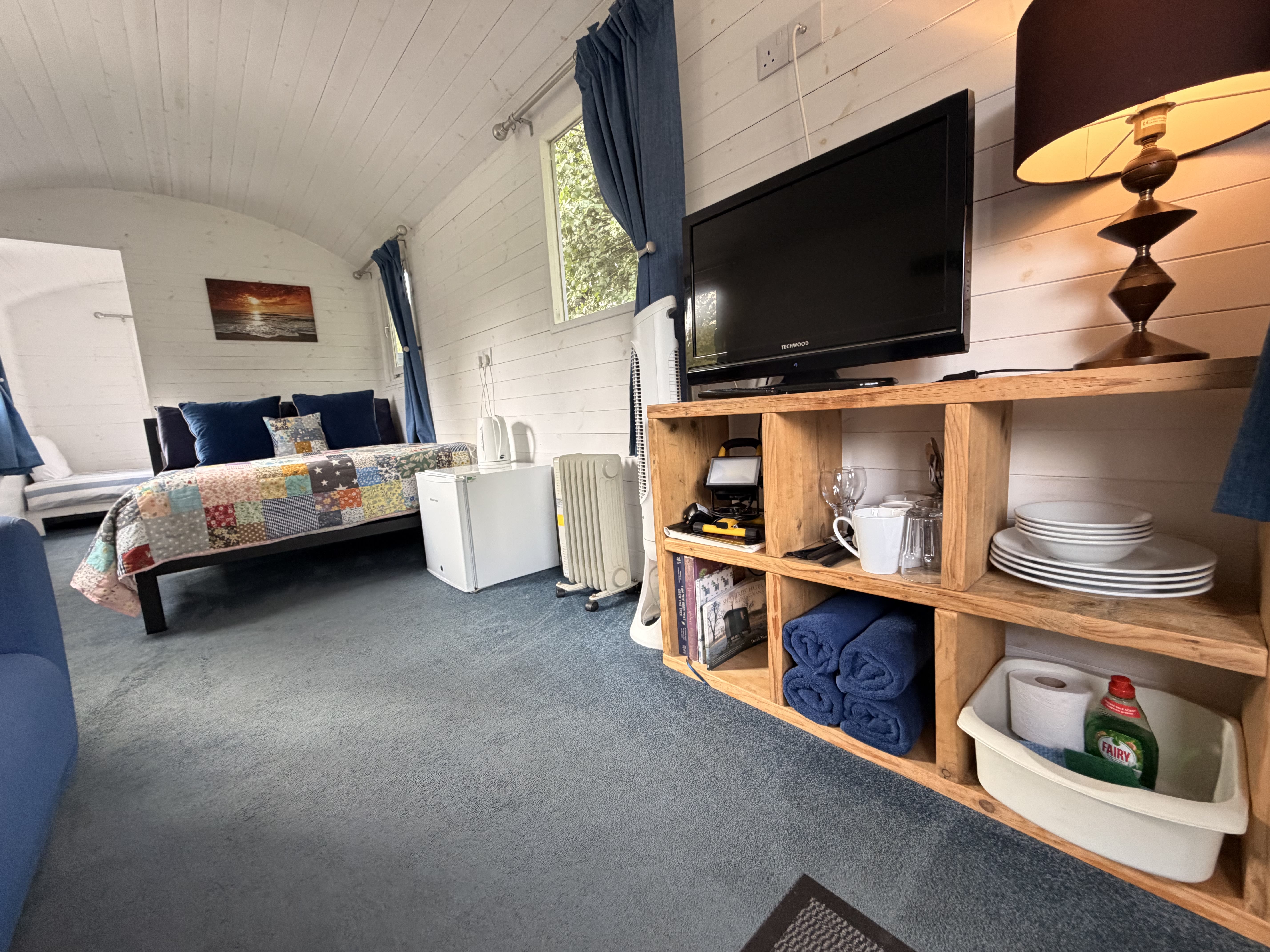 The kitchen corner: open wooden shelving with crockery, glasses and folded towels beside the TV, kettle, mini-fridge and a freestanding heater