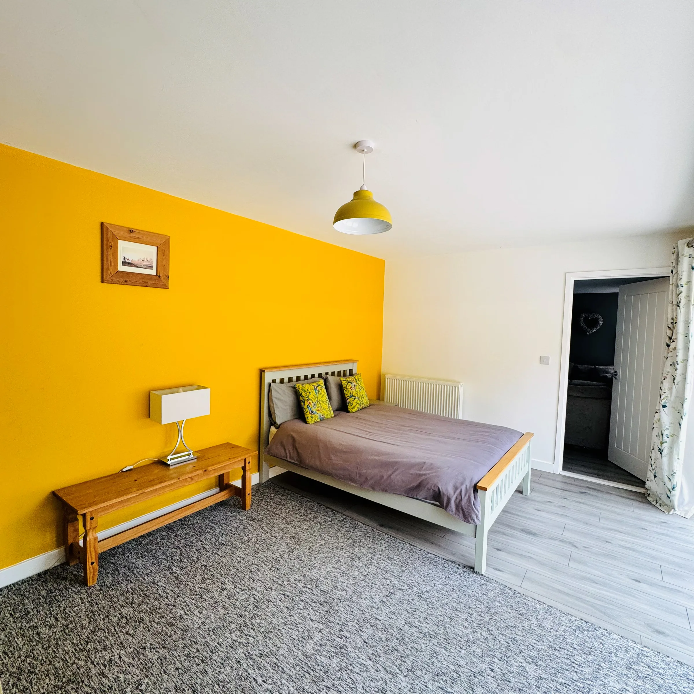 A single bedroom at Mordros with a mustard-yellow feature wall, a bench at the foot of the bed and an ensuite door beyond