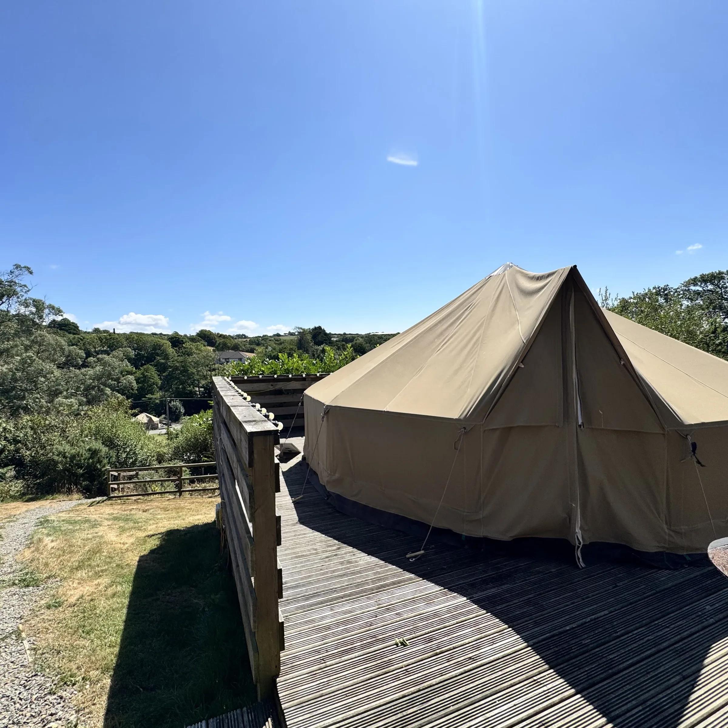 A sand-coloured canvas bell tent on a timber deck behind a slatted windbreak, guy ropes pegged out, looking over the treetops to the fields beyond