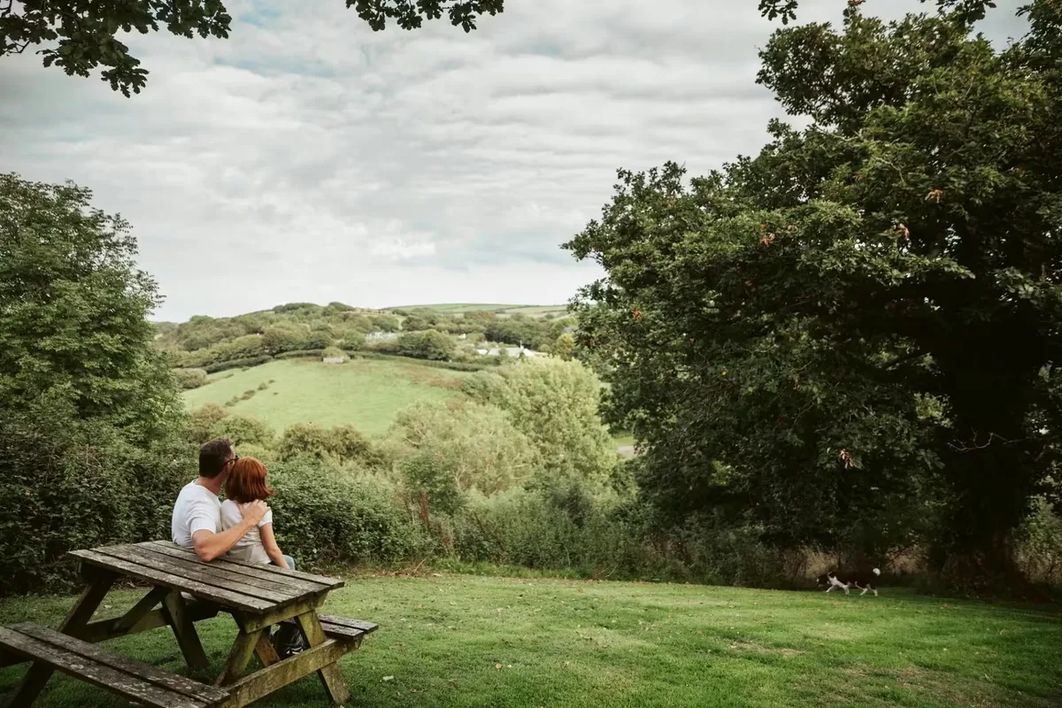 A couple sitting on a wooden picnic bench with their dog on the grass nearby, looking out across the green wooded valley
