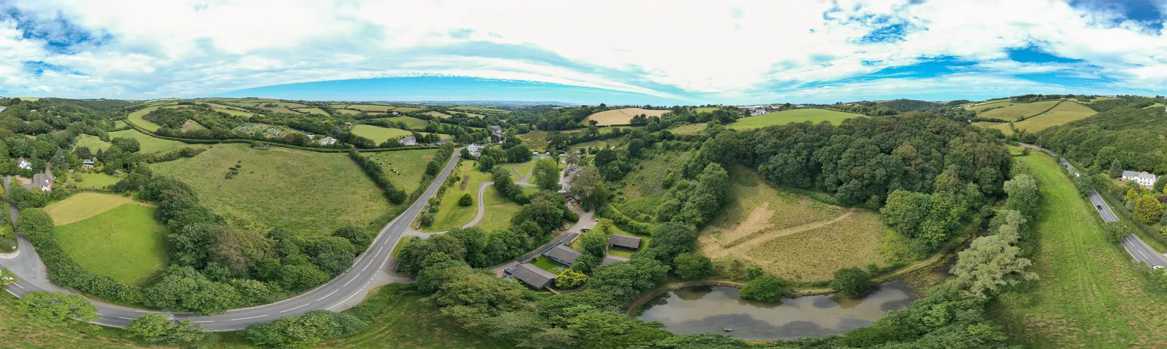 Aerial panorama of the wooded river valley, with pitches and lodges laid out along the fields and the North Cornwall coast beyond