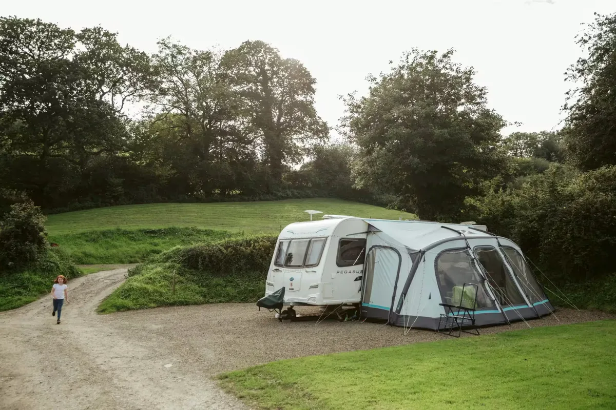 A caravan and its awning on a level gravel hardstanding pitch in the evening, with the track curving past and the field rising behind