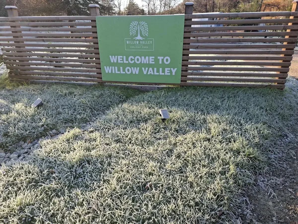 A green ‘Welcome to Willow Valley’ sign fixed to a slatted timber fence above frosted grass on a bright winter morning